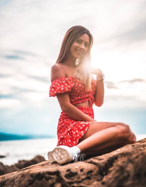 Zumaia, Gipuzkoa 'daki Itzurrun plajında kırmızı elbiseli esmer bir kadın. Bask Ülkesi. Yaşam tarzı oturumu, deniz kenarındaki bir kayanın üstünde.