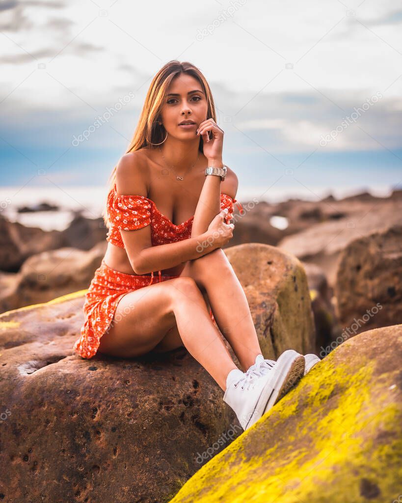 A young brunette Caucasian woman in a red dress on the beach of Itzurrun in the town of Zumaia, Gipuzkoa. Basque Country. Lifestyle session, sitting on a sea rock