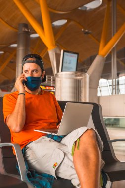 Koronavirüs salgınında hava yolculuğu, güvenli seyahat, sosyal mesafe, yeni normal. Yüzü maskeli genç bir adam kalkmayı beklerken bilgisayarla çalışıyor, havaalanları neredeyse boş.