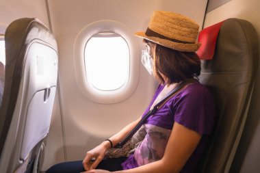 Koronavirüs salgınında hava yolculuğu, güvenli seyahat, sosyal mesafe, yeni normal. Bir uçağın içinde maskeli genç bir kadın uçarken pencereden dışarı bakıyor.