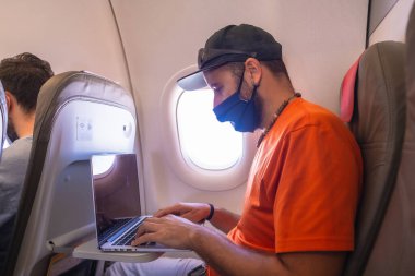 Koronavirüs salgınında hava yolculuğu, güvenli seyahat, sosyal mesafe, yeni normal. Bir uçağın içinde yüz maskesi olan genç bir adam bilgisayarla çalışıyor.