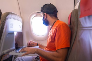 Koronavirüs salgınında hava yolculuğu, güvenli seyahat, sosyal mesafe, yeni normal. Bir uçağın içinde yüz maskesi olan genç bir adam bilgisayarla çalışıyor.