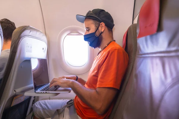 Air travel in the coronavirus pandemic, safe travel, social distance, new normal. A young man with a face mask inside an airplane working with a computer