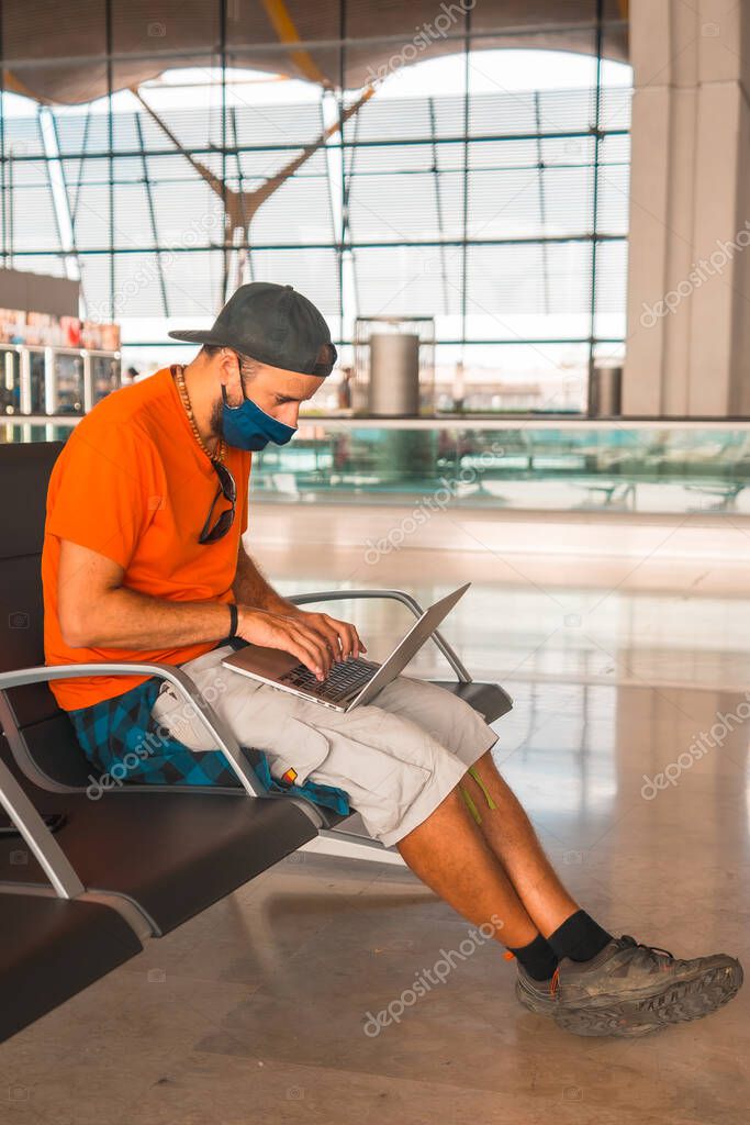 Air travel in the coronavirus pandemic, safe travel, social distance, new normal. A young man with a face mask working with the computer while waiting to take off, airports almost empty