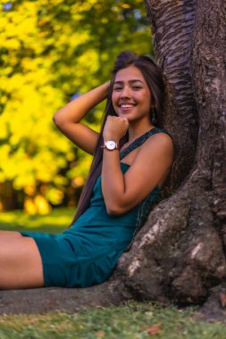 Yeşil elbiseli bir ağaca yaslanmış uzun düz saçlı, esmer, genç bir Latin. Parktaki bir ağacın yanındaki çimlerin üzerinde oturmuş eğleniyorum.