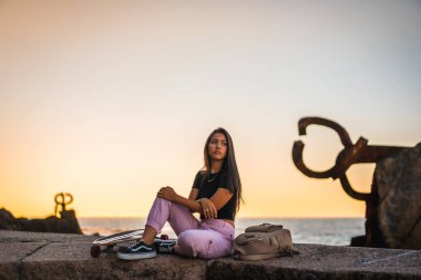 Siyah tişört ve pembe pantolonla kaykayın yanında oturan uzun düz saçlı esmer bir Latin. İspanya, San Sebatian 'da deniz kenarında bir heykelin yanında.