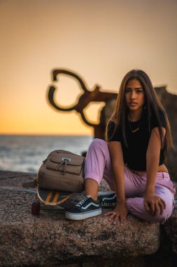 Siyah tişört ve pembe pantolonla kaykayın yanında oturan uzun düz saçlı esmer bir Latin. Günbatımında Peine del Viento adlı San Sebatian heykelinin yanında, İspanya