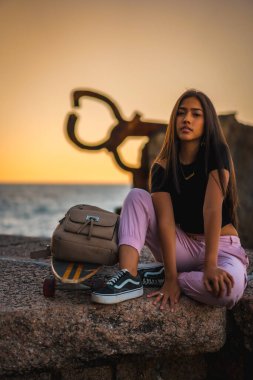 Siyah tişört ve pembe pantolonla kaykayın yanında oturan uzun düz saçlı esmer bir Latin. Günbatımında Peine del Viento adlı San Sebatian heykelinin yanında, İspanya