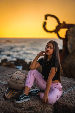 Siyah tişört ve pembe pantolonla kaykayın yanında oturan uzun düz saçlı esmer bir Latin. Günbatımında Peine del Viento adlı San Sebatian heykelinin yanında, İspanya