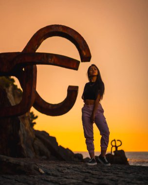 Uzun düz saçlı, kısa siyah tişörtlü ve pembe pantolonlu esmer bir Latin. Günbatımı, İspanya 'da Peine del Viento adlı San Sebatian heykelinin yanında duruyor.