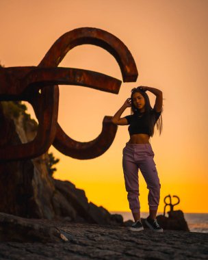 Uzun düz saçlı, kısa siyah tişörtlü ve pembe pantolonlu esmer bir Latin. Günbatımı, İspanya 'da Peine del Viento adlı San Sebatian heykelinin yanında duruyor.