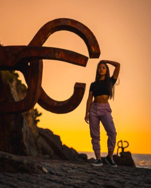 Uzun düz saçlı, kısa siyah tişörtlü ve pembe pantolonlu esmer bir Latin. San Sebatian 'ın ünlü heykeli Peine del Viento, İspanya' nın yanında turuncu gün batımı.