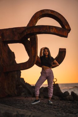 Uzun düz saçlı, kısa siyah tişörtlü ve pembe pantolonlu esmer bir Latin. İspanya 'da Peine del Viento adında ünlü bir Aziz Sebatian heykelinin yanına tünemiş.