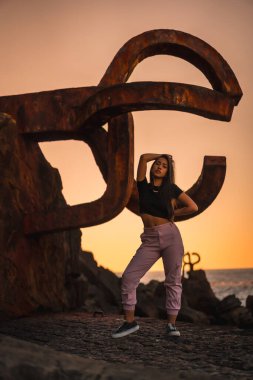 Uzun düz saçlı, kısa siyah tişörtlü ve pembe pantolonlu esmer bir Latin. İspanya 'da Peine del Viento adında ünlü bir Aziz Sebatian heykelinin yanına tünemiş.