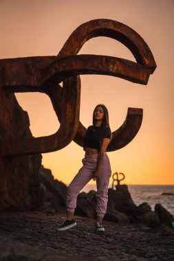 Uzun düz saçlı, kısa siyah tişörtlü ve pembe pantolonlu esmer bir Latin. İspanya 'da Peine del Viento adında ünlü bir Aziz Sebatian heykelinin yanına tünemiş.