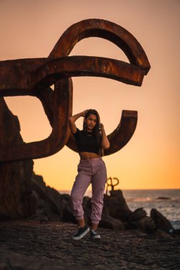 Uzun düz saçlı, kısa siyah tişörtlü ve pembe pantolonlu esmer bir Latin. İspanya 'da Peine del Viento adında ünlü bir Aziz Sebatian heykelinin yanına tünemiş.