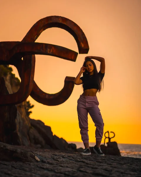 A young pretty brunette Latina with long straight hair in a short black T-shirt and pink pants. Orange sunset next to a famous sculpture of San Sebatian called Peine del Viento, Spain