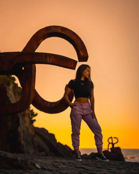A young pretty brunette Latina with long straight hair in a short black T-shirt and pink pants. Orange sunset next to a famous sculpture of San Sebatian called Peine del Viento, Spain