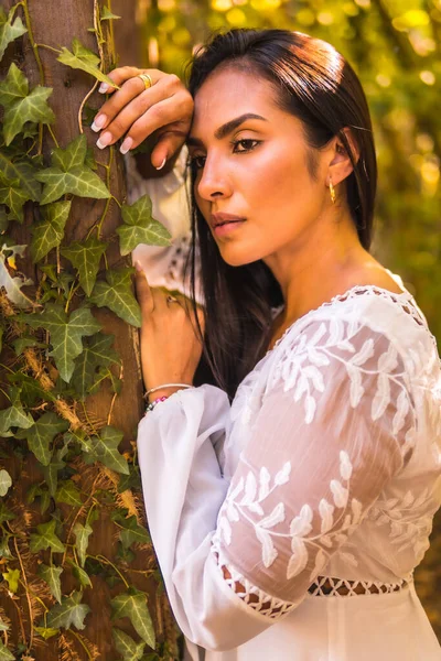 Lifestyle, brunette girl with straight hair in a beautiful white dress in a park resting in summer. Portrait of senxual look and innocent look leaning on a wood