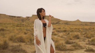 Yazın Bardenas Reales 'te beyaz kimonolu esmer bir manken ve siyah bir bikini pozu vermek. Yaşam tarzı, Navarra, İspanya. 4k video