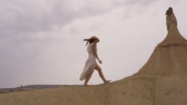 Beyaz elbiseli ve şapkalı beyaz tenli genç beyaz tenli Bardenas Reales 'teki Castildetierra' da yürüyor. Yaşam tarzı, Navarra, İspanya. 4k video