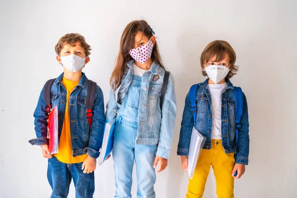 フェイスマスクをした3人の子供が学校に戻る準備ができています 新しい正常性 社会的距離 コロナウイルスパンデミック Covid 白い背景で学校に行くのを待って ペーストスペースをコピー — ストック写真