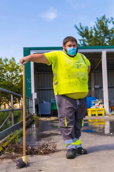 Worker in a recycling factory or clean point and garbage with a face mask and with security protections, new normal, coronavirus pandemic, covid-19. Portrait worker with a broom