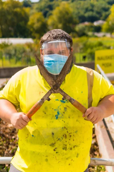 Worker in a recycling factory or clean point and garbage with a face mask and plastic protective screen, new normal, coronavirus pandemic, covid-19. Portrait with reflective vest and giant scissors