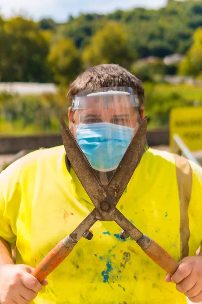 Worker in a recycling factory or clean point and garbage with a face mask and plastic protective screen, new normal, coronavirus pandemic, covid-19. Portrait with reflective vest and giant scissors