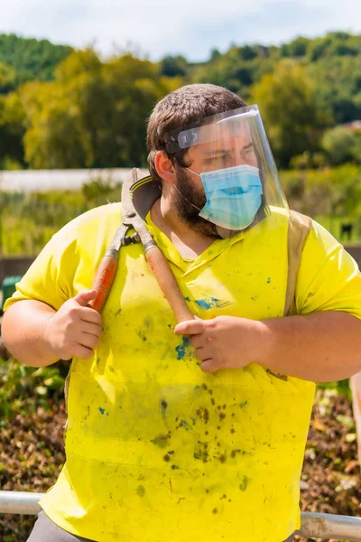Worker in a recycling factory or clean point and garbage with a face mask and plastic protective screen, new normal, coronavirus pandemic, covid-19. Portrait with reflective vest and giant scissors