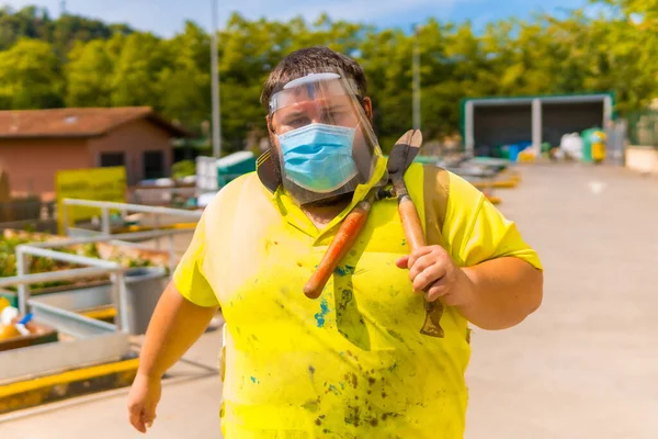 Worker in a recycling factory or clean point and garbage with a face mask and plastic protective screen, new normal, coronavirus pandemic, covid-19. Walking with reflective vest