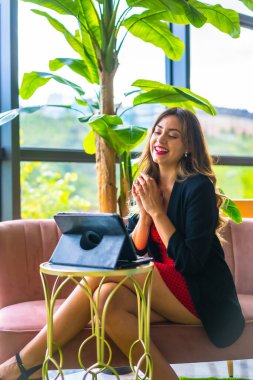 Sarışın, beyaz bir girişimci, iş görüşmesinin videosunu çekiyor, siyah ceket ve kırmızı elbise, evden işe giderken, yeni normal, pembe bir kanepede oturuyor ve oteldeki bazı yeşil bitkiler...