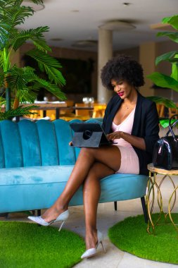 Siyah ırk ve afro saçlı genç girişimci bir iş görüşmesinin videosunu çekiyor, siyah ceket ve pembe elbise, tele-çalışma, yeni normal, mavi bir koltukta oturuyor ve oteldeki bazı yeşil bitkiler