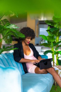 Siyah ırk ve afro saçlı genç girişimci bir iş görüşmesinin videosunu çekiyor, siyah ceket ve pembe elbise, tele-çalışma, yeni normal, mavi bir koltukta oturuyor ve oteldeki bazı yeşil bitkiler