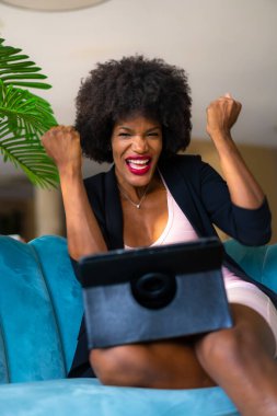 Siyah ırk ve afro saçlı genç girişimci büyük bir neşe ve şaşkınlıkla bir video görüşmesi yapıyor, tele-çalışma, yeni normal, mavi bir otelde bir kanepede oturuyor.