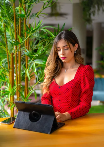 Sarışın, beyaz bir girişimci bir şehir otelinden iş videosu çekiyor. Tablet, kırmızı yönetici elbisesi, telekomünikasyon, otellerde çalışıyor.