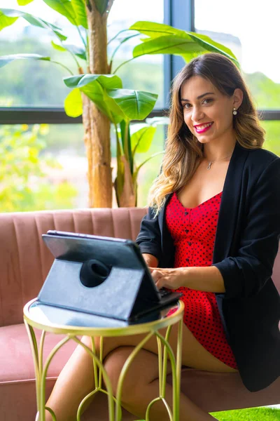 Genç sarışın, beyaz girişimci bir video konferansı düzenliyor siyah ceket ve kırmızı elbise, telekomünikasyon, yeni normal, pembe bir kanepede oturuyor.