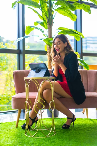 年轻的金发白种人企业家在工作会议上打了一个视频电话 穿着黑色夹克和红色衣服 电子通勤 新常态 坐在粉红色的沙发上和宾馆里的一些绿色植物 — 图库照片