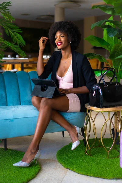 Siyah ırk ve afro saçlı genç girişimci bir iş görüşmesinin videosunu çekiyor, siyah ceket ve pembe elbise, tele-çalışma, yeni normal, mavi bir koltukta oturuyor ve oteldeki bazı yeşil bitkiler