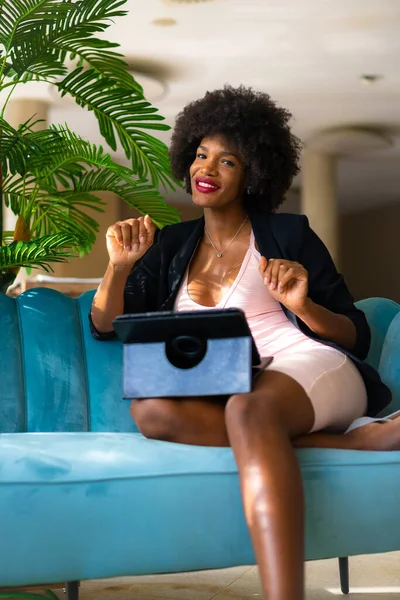 Siyah ırk ve afro saçlı genç girişimci bir iş görüşmesinin videosunu çekiyor, siyah ceket ve pembe elbise, tele-çalışma, yeni normal, mavi bir otelde bir kanepede oturuyor.