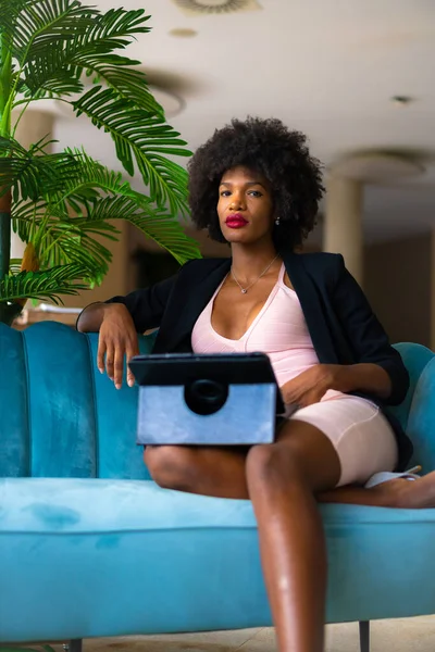 Siyah ırk ve afro saçlı genç girişimci bir iş görüşmesinin videosunu çekiyor, siyah ceket ve pembe elbise, tele-çalışma, yeni normal, mavi bir otelde bir kanepede oturuyor.