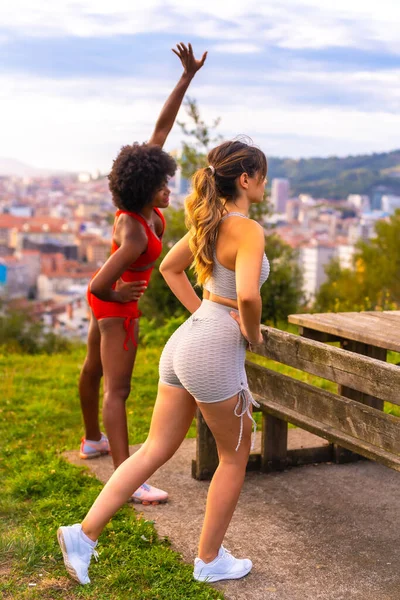 在公园开始运动之前 白人金发女孩和黑皮肤女孩都在做伸展运动 健康的生活 健康的身体 健康的女孩 灰色和红色的运动服 — 图库照片