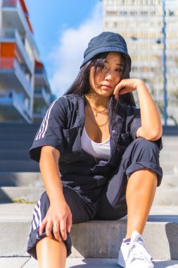 Siyah şapkalı ve siyah tulumlu genç Asyalı kız şehrin merdivenlerinde oturuyor. Şehirde, Çin uyruklu genç bir bayanla şehir toplantısı.