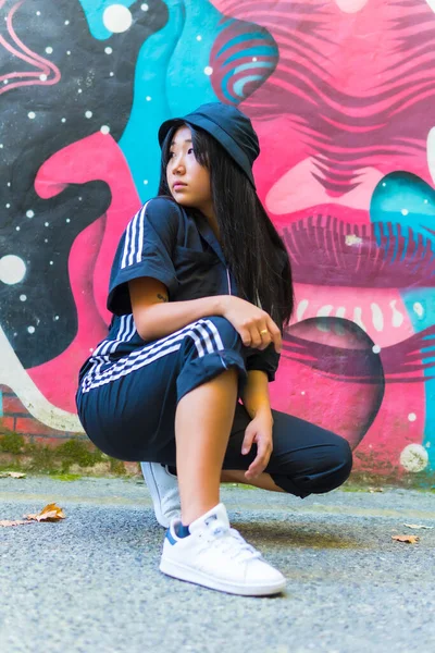 Young Asian rapper girl smiling in a black cap and black jumpsuit crouched on the ground in the city, photo trap pose. Urban session in the city with a young woman of Chinese nationality