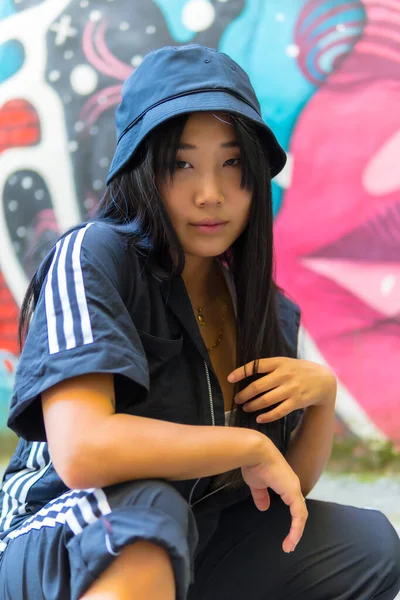 年轻的亚洲说唱歌手女孩 戴着黑色的帽子 穿着黑色的连衣裙蹲伏在城市的地面上 摆出一副照片陷阱的姿势 与一名中国裔青年妇女在城市举行的会议 — 图库照片