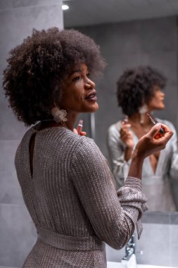 Afro saçlı genç bir zenci kız partiden önce gülümseyerek makyaj yapıyor. Özel parti, VIP partisi, gri gece elbisesi.