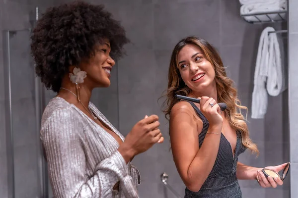 A young black girl with afro hair and a blonde Caucasian girl putting on makeup before the party with a smile. Exclusive party, vip party, gray and blue evening dress