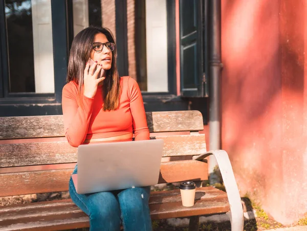 生活方式 年轻的高加索黑发姑娘穿着红色衬衫坐在公园里 一边喝着咖啡 和电脑一起工作 用手机打电话 — 图库照片