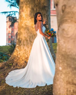 Uzun kuyruklu gelinlik giymiş esmer bir kız düğün kutlamasında bir ağacın yanındaki parkta.