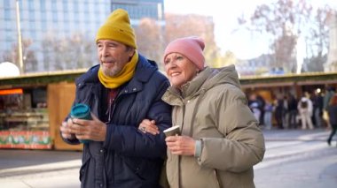Yaşlı çift kol kola yürüyor, sıcak içecekler içiyor ve kış pazarında Noel ruhunun tadını çıkarıyor.